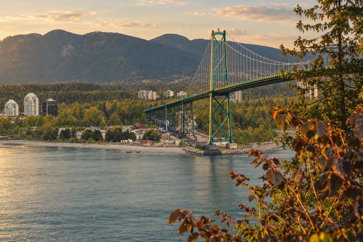 A green suspension bridge crossing calm water at sunset, with forested mountains behind.